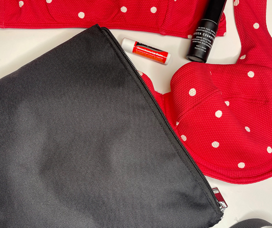 black bikini bag on a white background with red polka dot bikinis hanging out of it as well as sunscreen and chapstick