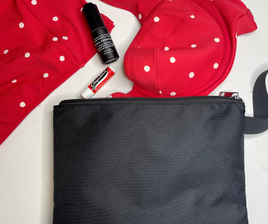 black bikini bag on a white background with red polka dot bikinis hanging out of it as well as sunscreen and chapstick
