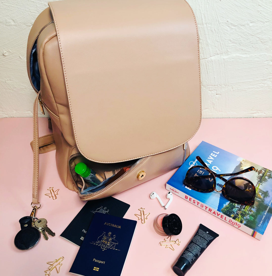 tan backpack against a pink backdrop and white brick wall. sunglasses, passports, makeup all sitting next to the backpack to show what to pack