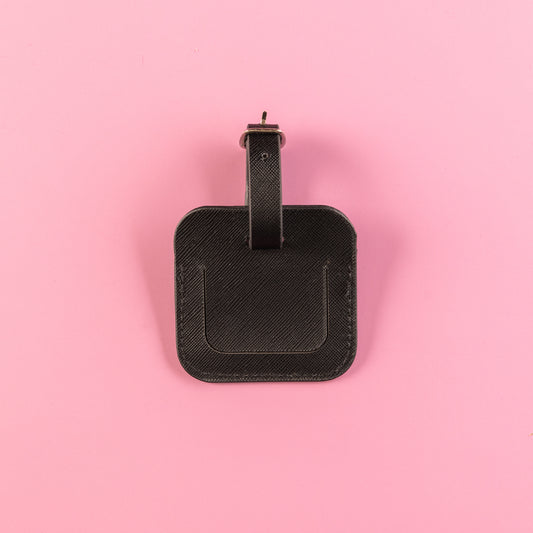 Black, square luggage tag with lift up flap to see owners details underneath. Luggage tag includes adjustable strap with silver hardware. All set on a pink background. This is the front of the luggage tag. 