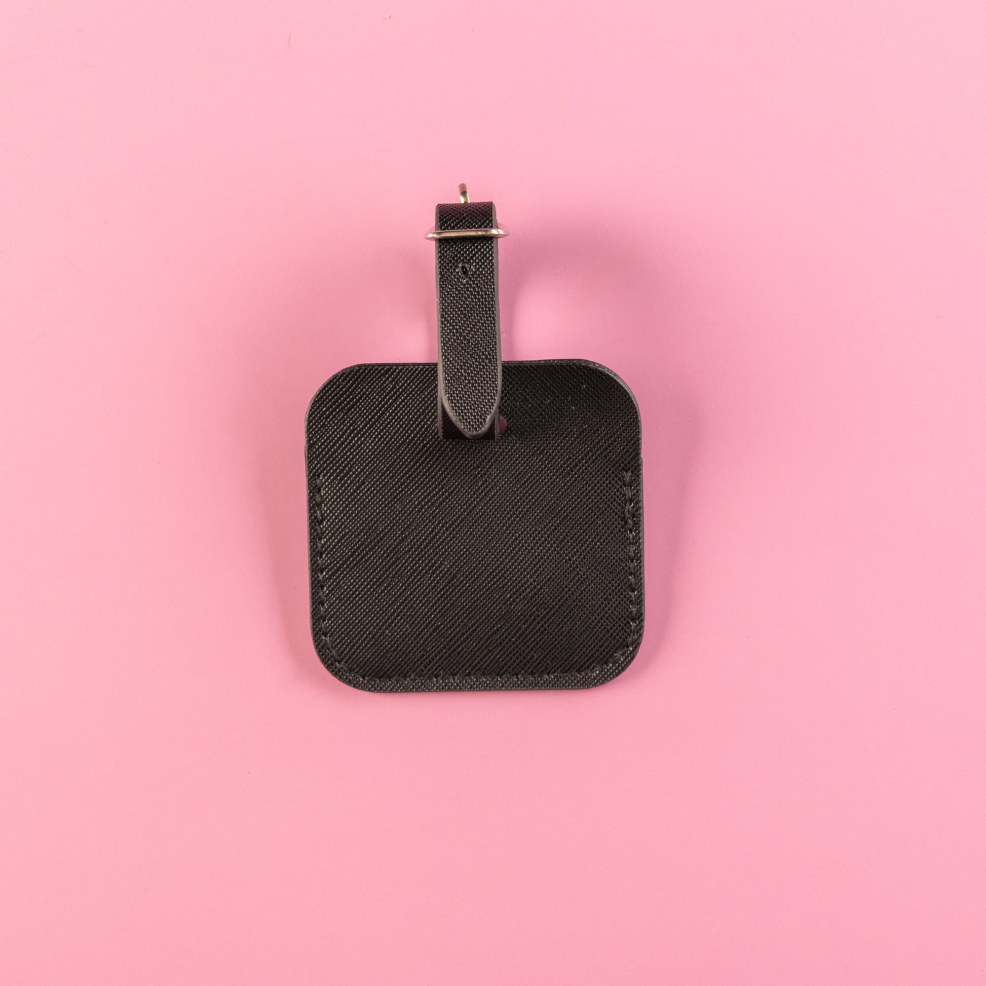 Black, square luggage tag with lift up flap to see owners details underneath. Luggage tag includes adjustable strap with silver hardware. All set on a pink background. This is the back of the luggage tag. 