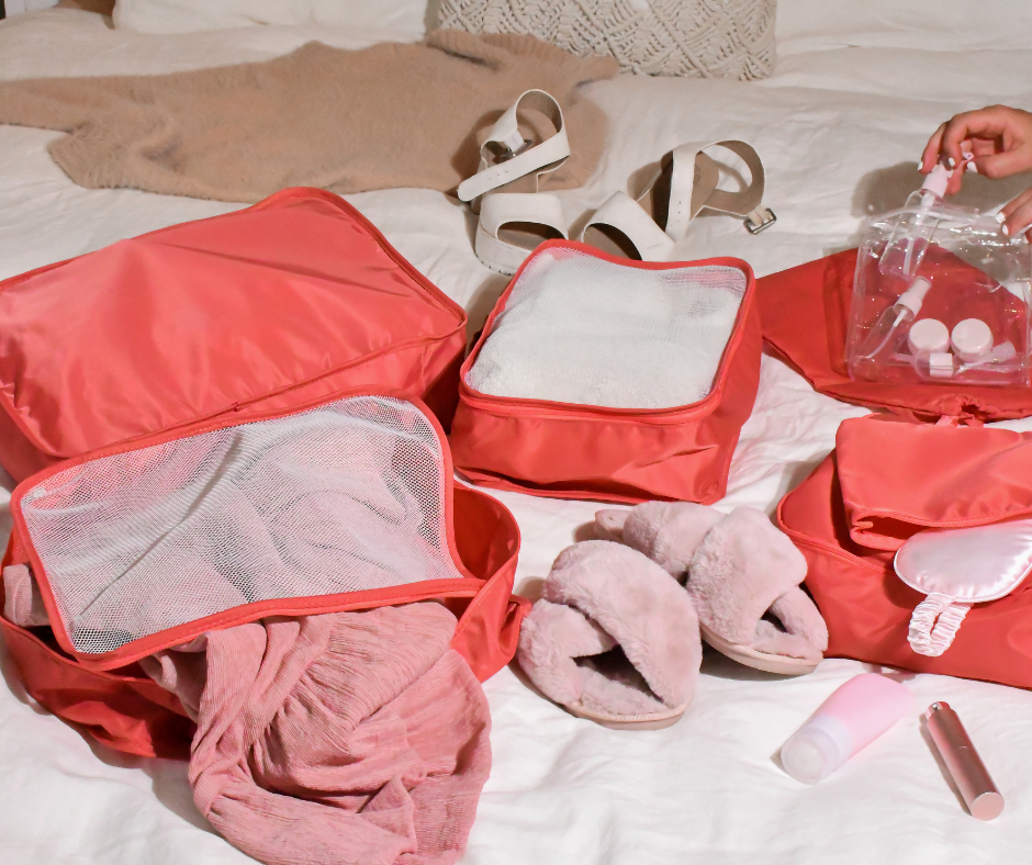 pink packing cubes and sleep/eye mask on a white bed cover with clothes spread out around them to show them being packed for a holiday