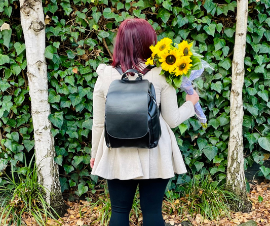 woman with black jeans and a grey coat on with the black backpack by midnight crusade on. standing against a green flower wall