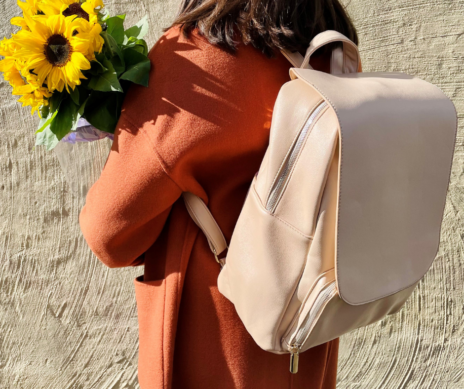 woman in an orange coat standing against a beige wall with the beige backpack from midnight crusade on. also holding sunflowers